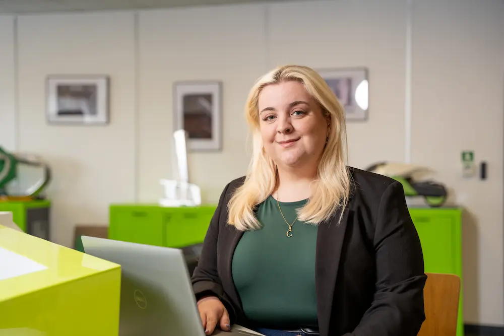 Charlotte Cooley sitting in front of a laptop at a counter with various items on display in the background.